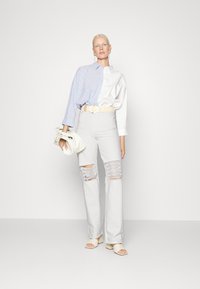 Camisa oversized azul clara e branca em riscas, jeans cinza claro de cintura alta com rasgos proeminentes nos joelhos, cinto bege e sandálias de slide bege.