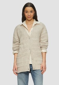 Beige gestrickte Strickjacke mit lockerer Passform, V-Ausschnitt, langen Ärmeln, Knopfverschluss und zwei vorderen Taschen, getragen über einem gestreiften Hemd.