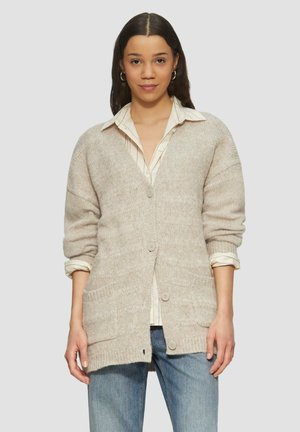 Cardigan en tricot beige avec une coupe décontractée, col en V, manches longues, fermeture à boutons et deux poches avant, porté sur une chemise rayée.
