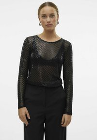 Schwarzes Netzoberteil mit langen Ärmeln, das ein Diamantmuster und einen runden Ausschnitt aufweist, über einem schwarzen Outfit getragen, mit glänzenden Akzenten und strukturiertem Stoff.