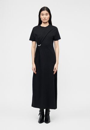 Femme aux longs cheveux foncés portant une robe midi noire à manches courtes avec une taille froncée et une broche argentée, associée à des bottines noires.