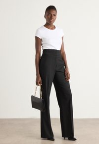 Un haut blanc à manches courtes, un pantalon noir taille haute à jambes larges et un sac à main noir avec une bandoulière en chaine dorée. Texture lisse, design minimaliste.