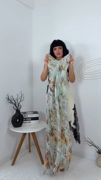 Žena s crnom kosom drži lepršavu, višebojnu haljinu u bijeloj prostoriji sa malim okruglim stolom i dekorativnim granama.