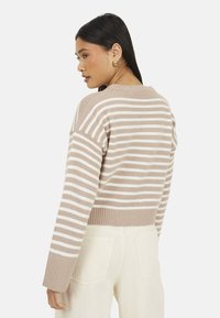 Donna con lunghi capelli scuri, che indossa orecchini a cerchio dorati, un maglione a righe beige e bianche e pantaloni bianco sporco, rivolta di lato con la testa girata.
