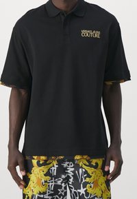Czarny polo z krótkimi rękawami, z logo Versace Jeans Couture w kolorze złotym oraz wzorzystymi wykończeniami na mankietach. Wykonany z bawełny, w luźnym fasonie.