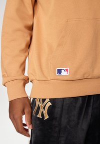 Sweat-shirt kaki avec des poignets côtelés et une poche avant, arborant un logo MLB bleu et rouge. Associé à un pantalon en velours noir avec des détails dorés.