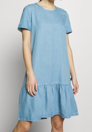 Femme portant une robe en denim bleu clair à manches courtes avec un ourlet froncé, debout devant un fond clair uni.