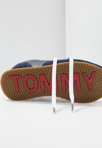 Basket bleue en suède avec une semelle en caoutchouc, arborant un logo rouge "TOMMY" et des lacets blancs. Texture en chevrons sur la semelle extérieure.