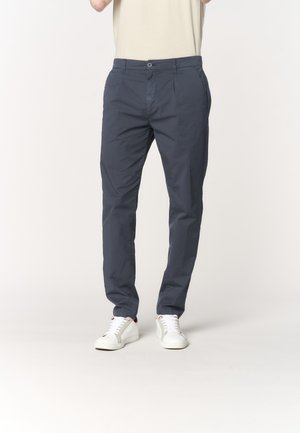 Uomo con chinos blu navy e sneakers bianche, in piedi contro uno sfondo chiaro e uniforme.