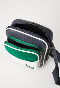 Piccolo borsello a tracolla in pelle blu navy con una tasca frontale con zip bianca e verde. Caratterizzato da doppie zip e una tracolla staccabile.