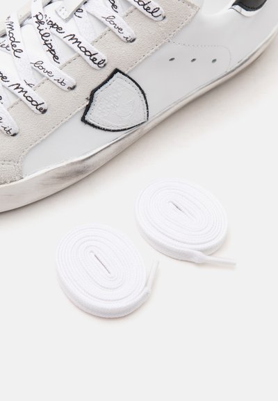 Baskets en cuir blanc avec des accents noirs et beiges, lacets de marque avec texte manuscrit et deux lacets blancs enroulés à côté.