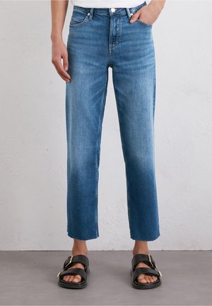 Rechtvallende blauwe spijkerbroek met lichte vervaging, gedragen met zwarte sandalen met gouden gespen, bij een persoon die staat tegen een neutrale muur.