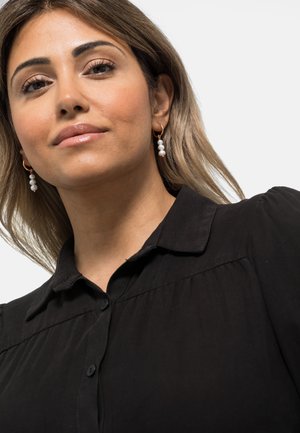 Schwarze Bluse mit einem geknöpften Kragen und Knopfleiste, die über Faltendetails an den Schultern verfügt. Goldene Creolenohrringe mit drei weißen Perlen.