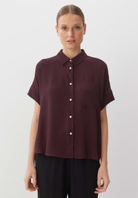 Burgundy short-sleeve shirt with a collar, front pocket, and button closure made from a smooth fabric. Features a relaxed fit.