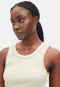 Lichtbeige ribbelige tanktop met witte rand, met een ronde hals. Zilveren ketting met een cilindrische hanger. Donker gevlochten haar.