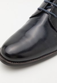 Chaussure habillée en cuir noir poli avec des lacets foncés sur un fond blanc uni, montrant les détails avant et latéraux.