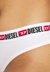 String en coton blanc avec une ceinture élastique noire et rouge marquée "DIESEL". Texture lisse et design ajusté.