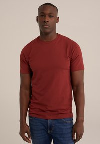 Rde majica iz bombaža s kratkimi rokavi in okroglo ovratnikom, v kombinaciji z modrimi jeans hlačami. Tkanina se zdi gladka in lahka.