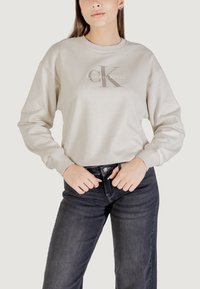 Sudadera beige con cuello redondo y mangas largas, que presenta un gráfico frontal con "Calvin Klein" en letras metálicas.