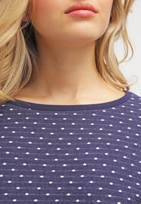 Blondynka w fioletowej dzianinowej bluzce w białe kropki, eksponująca szyję, podbródek, usta i część włosów.