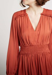 Robe couleur rouille avec un décolleté en V, des manches longues, des accents d'épaules froncés et une taille smockée. Tissu lisse avec une jupe fluide.