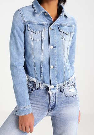 Frau trägt eine hellblaue, ausgewaschene, kurze Jeansjacke mit silbernen Knöpfen und passende hoch taillierte, ausgewaschene Jeans.