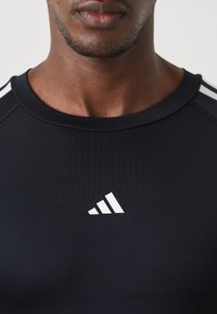 Fekete sportpóló kerek nyakkivágással, sima, rugalmas anyagból készült. Három fehér csíkkal díszített a vállakon, és fehér logo található rajta.