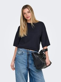 Mlada ženska z dolgimi blond lasmi, oblečena v črno preveliko majico in ohlapne modre jeans hlače, drži črno usnjeno torbico za ročaj.