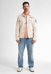 Chaqueta de mezclilla beige con cuello puntiagudo, dos bolsillos en el pecho y un parche circular en el brazo izquierdo. Lleva puesta una camisa rosa clara y jeans azules.