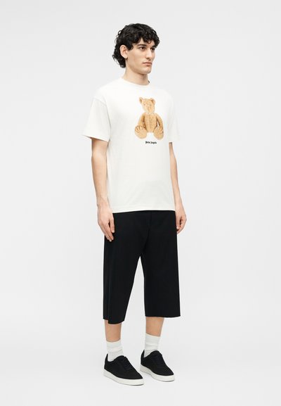 Jeune homme aux cheveux bouclés portant un t-shirt blanc avec un ours en peluche, un short noir au niveau des genoux, des chaussettes blanches et des baskets noires.