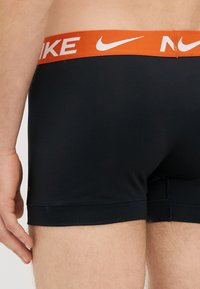 Μαύρα μποξεράκια με πορτοκαλί ζώνη Nike. Ο ύφασμα φαίνεται λεία και ελαστική, με σχεδίαση που αγκαλιάζει το σώμα και κοντό μήκος ποδιού.