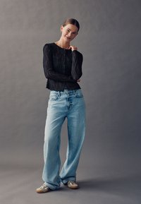 Schwarzes, transparentes Langarmhemd mit vertikalen Streifen, kombiniert mit hellblauen Jeans mit weitem Bein. Das Modell trägt helle Sneaker. Schlichter Hintergrund.