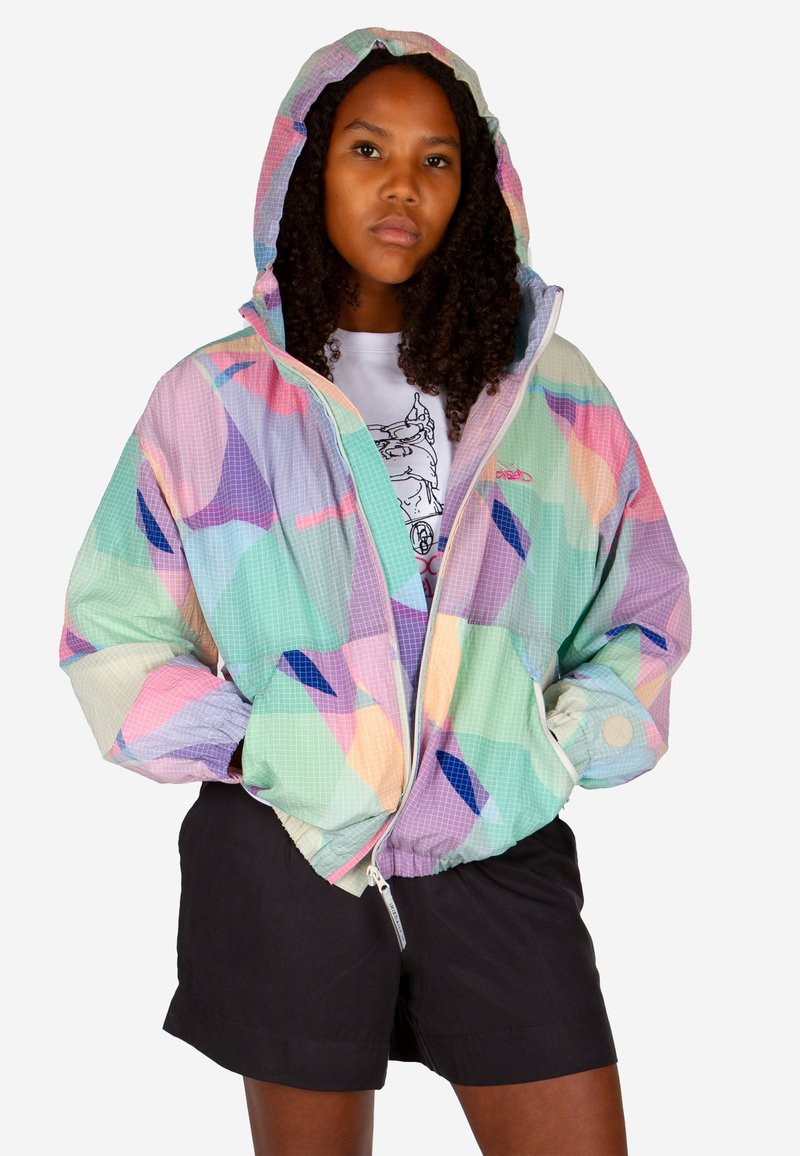 Woman with curly hair wearing a multicolored pastel hooded jacket, white graphic tee, and black shorts, standing with hands in jacket pockets.