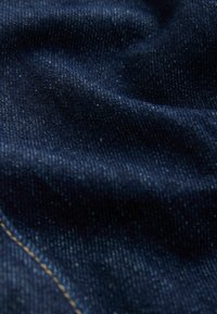 Tissu en denim bleu foncé avec une texture diagonale visible et une seule ligne de couture beige dans le coin inférieur gauche.