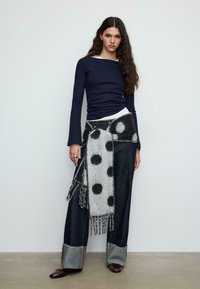 Temno modra majica z dolgimi rokavi, sivo-črni pikeš šal, široke kavbojke z raztrganimi robovi in rjavi čevlji.