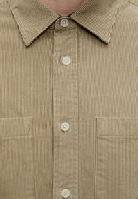 Chemise beige en velours côtelé avec col et deux poches poitrine, partiellement boutonnée.