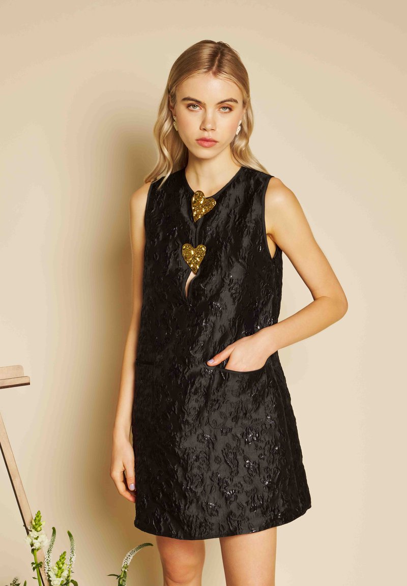 Młoda kobieta o blond włosach, ubrana w bezrękawnikową, czarną, teksturowaną sukienkę z dwoma złotymi guzikami w kształcie serc, stojąca z jedną ręką w kieszeni.