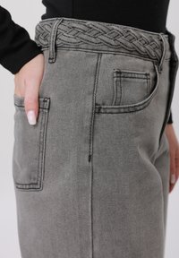 Nærbilde av en person som har på seg grå denimbukser med et flettet stoffbelte, hånden hvilende i baklommen, og en svart genser med lange ermer.