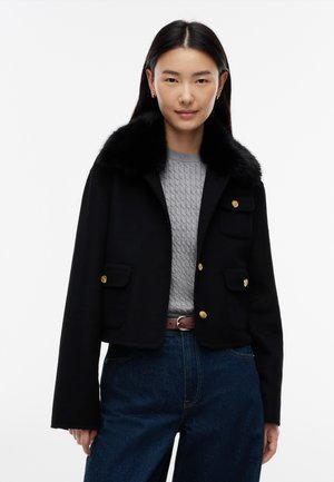 Femme portant une veste courte noire avec col en fourrure, un pull en maille gris, un jean bleu et une ceinture marron, debout devant un fond blanc.