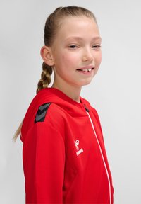 Rød zip-up hættetrøje med en tekstureret stof, der har en sort detalje på skulderen og et hvidt logo. Glat finish og tætsiddende design.