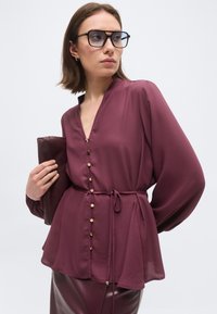 Γυναίκα που φοράει μαρουν κοντομάνικο πουκάμισο με χρυσά κουμπιά και δέσιμο στη μέση, κρατώντας τσαντάκι που ταιριάζει και φοράει μεγάλα μαύρα γυαλιά με πλαίσιο.