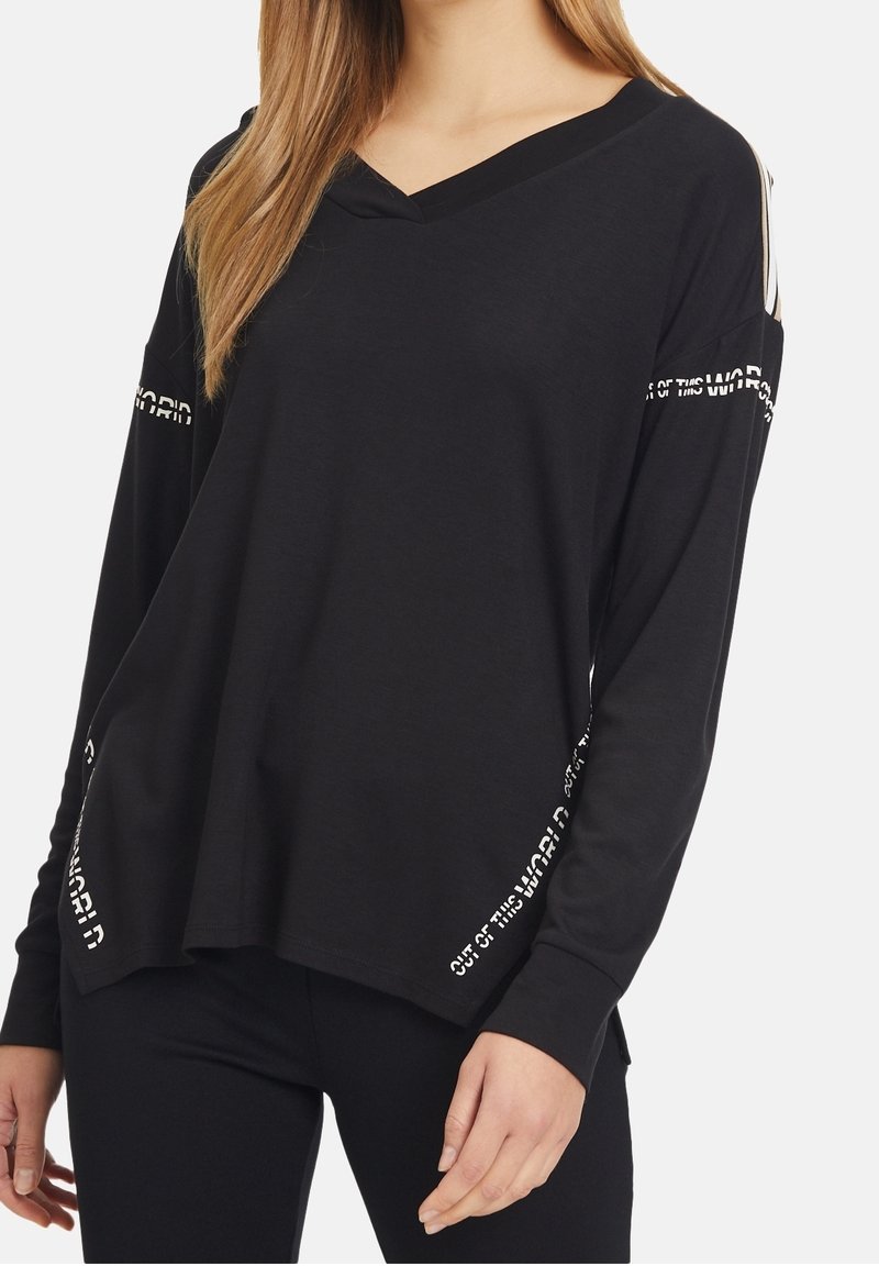 Femme portant une chemise noire à manches longues et ample avec un texte blanc le long des manches et des côtés indiquant "OUT OF THIS WORLD."