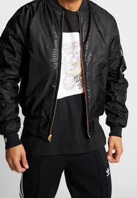 Svart bomberjacka med en glänsande yta, dragkedja fram, ribbade ärmslut och sidofickor. Buren över en svart t-shirt med grafisk tryck och svarta byxor.