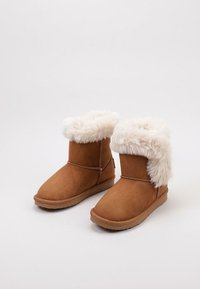 Botas de tobillo de ante color beige con puño de piel sintética blanca, punta redondeada y suela de goma plana, que presentan costuras decorativas a lo largo de la costura.