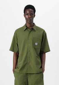 Chemise verte à manches courtes en tissu texturé. Présente une poche poitrine avec un logo et une coupe décontractée. Design minimaliste avec fermeture à boutons.