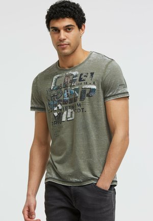 Mann mit lockigem schwarzen Haar trägt ein verblasstes grünes Grafik-T-Shirt und dunkle Jeans, steht mit einer Hand in der Tasche vor einfachem Hintergrund.