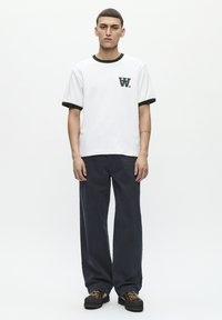 White cotton t-shirt with green ribbed collar and cuffs, featuring a small green logo. Worn with dark navy wide-leg trousers and brown shoes.