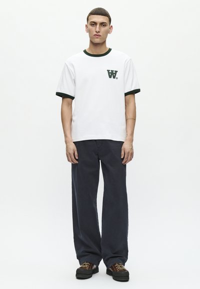 T-shirt bianco in cotone con colletto e polsini a coste verdi, con un piccolo logo verde. Indossato con pantaloni ampi blu scuro e scarpe marroni.