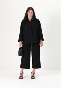 Must kollane villasegu mantli, millel on krae ja kolm nupu, koos mustade laiade pükstega ja rihmadega mustade sandaalidega, hoides peos väikest musta kotti.