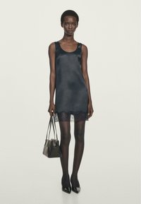 Black satin slip dress with lace hem, sleeveless design, worn with black sheer tights and pointed toe heels, held with a small black handbag.